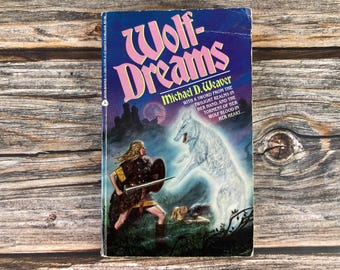 May include: A paperback book cover with a blue background and the title "Wolf-Dreams" in pink lettering. The author's name, "Michael D. Weaver", is printed below the title. The cover features an illustration of a woman with a sword and a wolf-like creature.