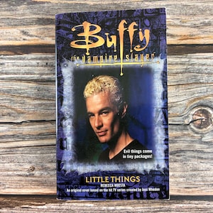 May include: A paperback book titled Buffy the Vampire Slayer: Little Things by Rebecca Moesta. The cover features a portrait of a blonde man against a dark blue background, with the text 'Evil things come in tiny packages!' printed below.