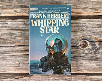 May include: A vintage paperback book titled "Whipping Star" by Frank Herbert. The cover features a blue background with a large, spherical object atop a rocky formation, with the ocean in the background. The book is a Berkley Medallion Book.