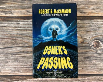 May include: A paperback book titled "Usher's Passing" by Robert R. McCammon. The cover features a dark blue background with a large, glowing wolf's face and a figure on a hill. The title is in yellow.