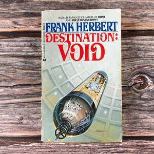 May include: A vintage paperback book titled "DESTINATION: VOID" by Frank Herbert. The cover features a futuristic illustration of a spacecraft and the title in large red letters. The book is set against a wooden background.