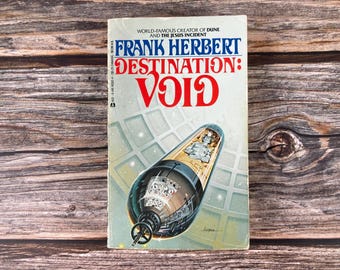 May include: A vintage paperback book titled "DESTINATION: VOID" by Frank Herbert. The cover features a futuristic illustration of a spacecraft and the title in large red letters. The book is set against a wooden background.
