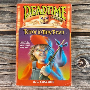 May include: A paperback book cover with a red background and the title "Deadtime Stories: Terror in Tiny Town". The cover features an illustration of a young boy with a red cap and a sinister expression, holding a stick and a sack. The text "Hurley's the leader of the pack. A very nasty pack." is printed below the illustration.