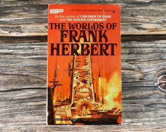 May include: A book cover with an orange background and a futuristic city scene. The title is "The Worlds of Frank Herbert" and the book is by the author of "Children of Dune" and "The Dosadi Experiment".