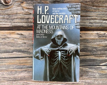 May include: A paperback book cover featuring a spooky illustration of a hooded figure with a skeletal ribcage visible beneath their cloak. The book title is "At the Mountains of Madness" by H.P. Lovecraft. The subtitle is "and Other Tales of Terror".