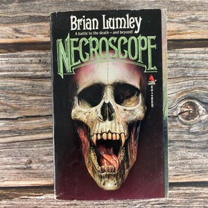 May include: A paperback book titled "Necroscope" by Brian Lumley. The cover features a detailed illustration of a skull with an open mouth and exposed tongue. The title is in green, and the background is black.