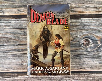 May include: A vintage paperback book titled "Demon Blade" with fantasy artwork. The cover features a woman with a sword, a skeletal figure, and a creature. The authors' names are Mark A. Garland and Charles G. McGraw.