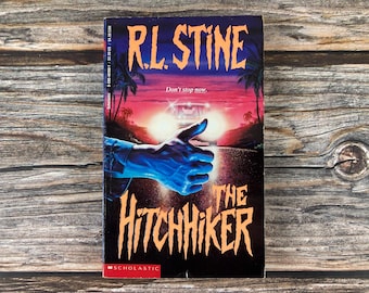 Puede incluir: Un libro de bolsillo titulado "The Hitchhiker" de R.L. Stine. La portada presenta una mano azul haciendo autostop, con los faros de un coche al fondo. El título es naranja sobre un fondo oscuro.