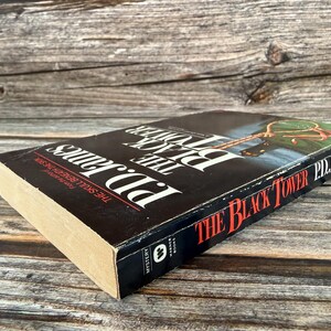 May include: A hardcover book titled "The Black Tower" by P.D. James. The book has a black cover with white and red text. The spine shows the title and author's name. The book is lying on a wooden surface.
