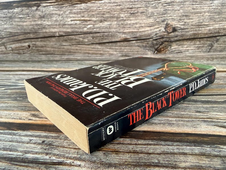May include: A hardcover book titled "The Black Tower" by P.D. James. The book has a black cover with white and red text. The spine shows the title and author's name. The book is lying on a wooden surface.