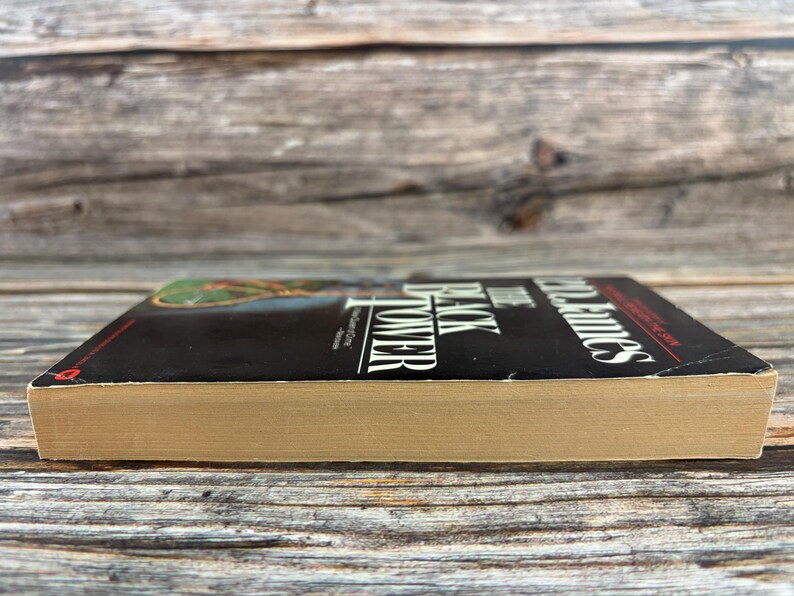 May include: A hardcover book with a black cover and the title "The Black Tower" by P.D. James. The book's pages are a light tan color. The book is set against a weathered wooden background.