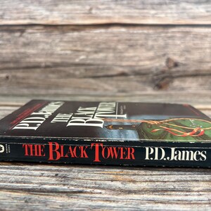 May include: A hardcover book titled "The Black Tower" by P.D. James. The book has a black cover with red and white text. The cover art features a green and gold illustration. The book is lying on a wooden surface.