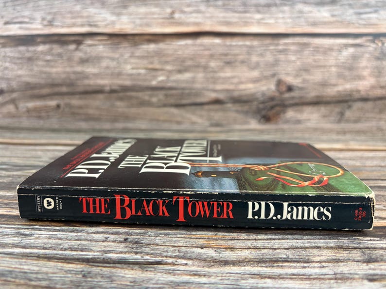 May include: A hardcover book titled "The Black Tower" by P.D. James. The book has a black cover with red and white text. The cover art features a green and gold illustration. The book is lying on a wooden surface.