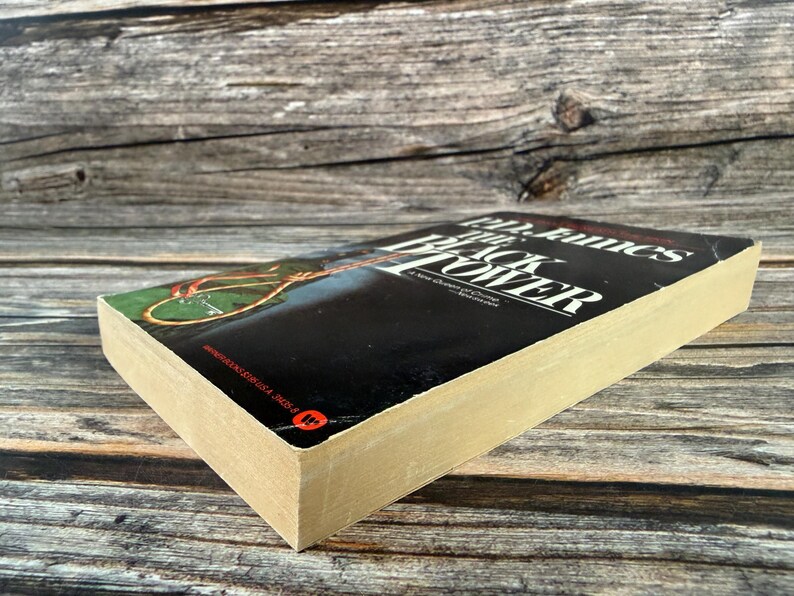 May include: A hardcover book titled "The Black Tower" by PD James. The book has a black cover with white text and a colorful illustration. The pages are yellowed with age, and the book is resting on a wooden surface.