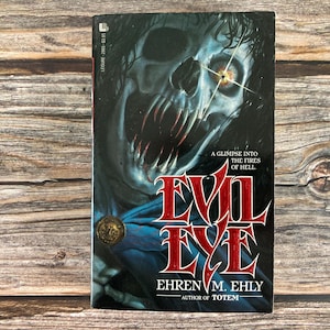 May include: A paperback book titled "Evil Eye" by Ehren M. Ehly. The cover features a skull-like figure with sharp teeth and a glowing eye, set against a dark background. The book's title is in large, red, stylized letters.