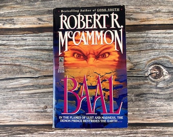 May include: A paperback book titled "BAAL" by Robert R. McCammon. The cover features a demonic face with fiery eyes and the text "IN THE FLAMES OF LUST AND MADNESS, THE DEMON PRINCE BESTRIDES THE EARTH". The book is set against a wooden background.