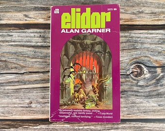 May include: A vintage paperback book cover with a purple background and the title "Elidor" in green letters. The author's name, "Alan Garner", is printed below the title in white letters. The cover features an illustration of a dark, mysterious doorway with a creature inside.