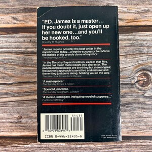 May include: A black paperback book cover with white text and a barcode. The cover features quotes about the author P.D. James. The book's ISBN is 0-446-31435-8. The background is a wooden surface.