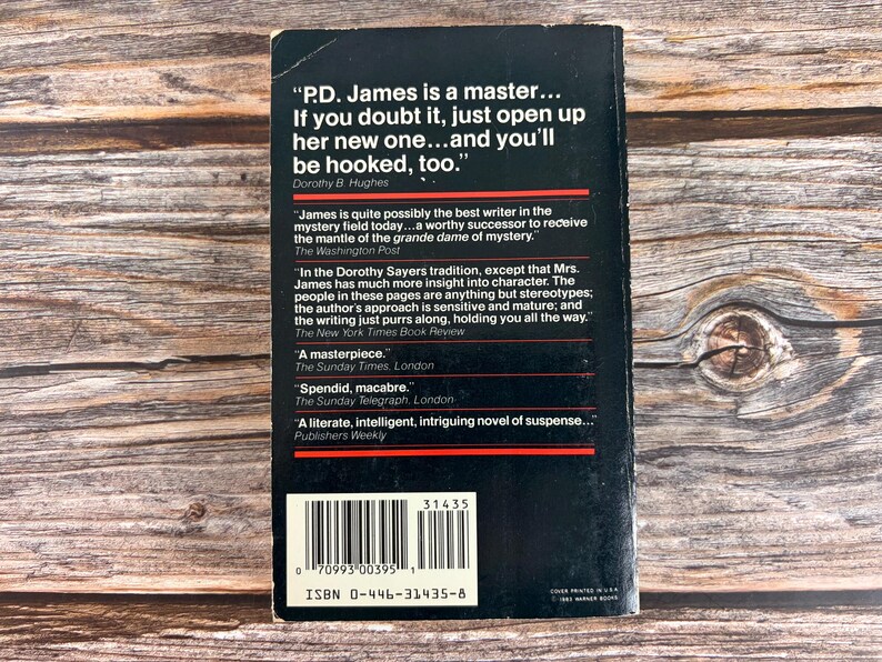 May include: A black paperback book cover with white text and a barcode. The cover features quotes about the author P.D. James. The book's ISBN is 0-446-31435-8. The background is a wooden surface.