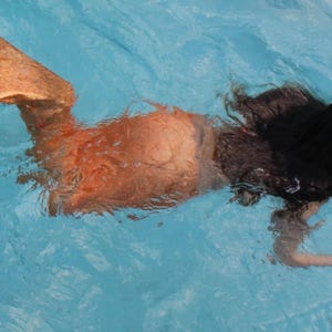 Puede incluir: Una persona con una cola de sirena nada en una piscina azul. La cola es naranja y el pelo de la persona es largo y oscuro.