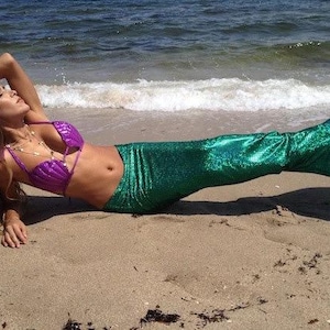 May include: A woman wearing a purple seashell bikini top and a green mermaid tail on a sandy beach.