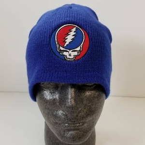 May include: A blue knit beanie featuring an embroidered patch with a skull and lightning bolt design in red, white, and blue. The beanie is displayed on a mannequin head. This hat is a music-themed accessory.