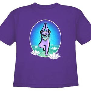 May include: Purple t-shirt with a graphic of a cartoon bear in a yoga pose. The bear is surrounded by lotus flowers and a blue and green background.