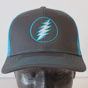 May include: A black and teal trucker hat with a white lightning bolt logo in a teal circle. The logo is embroidered on the front of the hat.