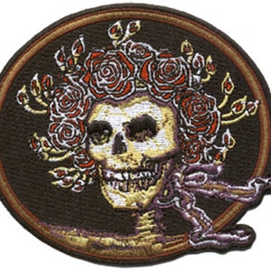 May include: A circular embroidered patch with a brown background featuring a white skull with a crown of red roses. The skull has a gold outline and the patch has a gold border.