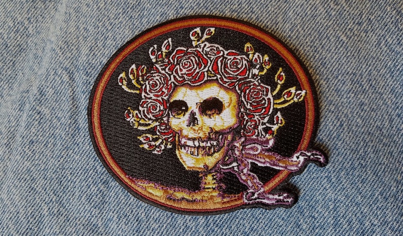 May include: A black and gold embroidered patch featuring a skull with a crown of red roses. The skull is surrounded by a gold circle.