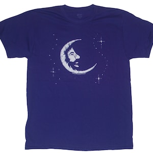May include: A purple t-shirt with a white graphic of a crescent moon with a face inside. The moon is surrounded by small white stars.