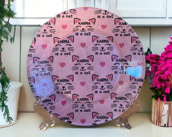 May include: A pink glass plate with a repeating pattern of black cat faces and the text "Karma is a cat". The plate is on a gold metal stand.