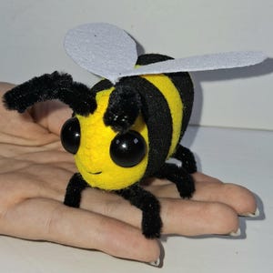 May include: A small bee figurine crafted from yellow and black fuzzy material with large, round, black plastic eyes. The bee features two white felt wings on its back and thin, black, pipe-cleaner-style legs and antennae extending from its body.