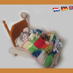 Op de afbeelding: Een gehaakt bed met een patchwork-deken en verschillende gehaakte figuren die onder de dekens slapen. De figuren omvatten een man, een vrouw, een kind, een hond en een kat.