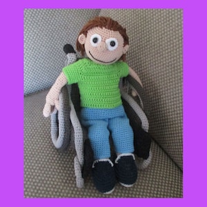 Może przedstawiać: Przędziona lalka z brązowymi włosami, zieloną koszulą, niebieskimi spodniami i czarnymi butami siedzi w przędzonej wózku inwalidzkim. Lalka ma duże, białe oczy i uśmiechniętą twarz.