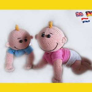 Op de afbeelding: Twee gehaakte babypoppen, een in roze en een in blauw, kruipen op een wit oppervlak. De poppen hebben bruine ogen en glimlachende gezichten.