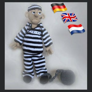 May include: A crocheted prisoner doll wearing a black and white striped jumpsuit with the number "12-41" on the chest. The doll is holding a crocheted gray ball and chain. The background features flags of Germany, the United Kingdom, and the Netherlands.