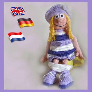 Könnte beinhalten: Eine gehäkelte Puppe, die ein lila-weiß gestreiftes Kleid, eine lila Baskenmütze mit einer weißen Blume und lila Schuhe trägt. Die Puppe sitzt auf einer gelben Toilette.