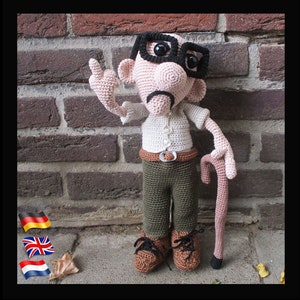 Op de afbeelding: Een gehaakte pop met een wit shirt, groene broek en een bruine stok. De pop draagt een bril en heeft een snor. De pop staat voor een bakstenen muur.