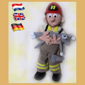 Pode incluir: Uma boneca de bombeiro de crochê vestindo um capacete vermelho com uma faixa amarela e segurando uma boneca de gato de crochê cinza.
