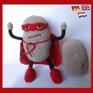 Op de afbeelding: Een gehaakt aardappel speelgoed dat een rode cape en een rood masker met gele ogen draagt. De aardappel staat met zijn armen omhoog in een superheldenpose. Er ligt een tweede gehaakt aardappel speelgoed op de grond.