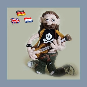 May include: A crocheted doll with long brown hair, wearing a black shirt with a skull and crossbones design, green pants, and holding a brown guitar. The doll is wearing a black wristband and has a white and brown hat.