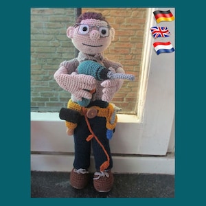 Op de afbeelding: Een gehaakte pop met een bril en een gereedschapsgordel met een boormachine in zijn handen. De pop draagt ​​bruine schoenen en een blauwe broek.