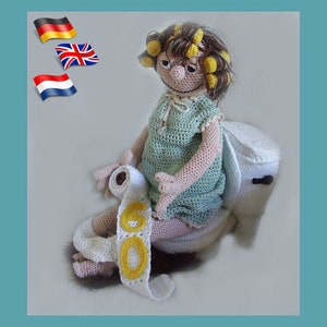 May include: A crocheted doll with brown hair and yellow curlers sits on a white crocheted toilet. The doll is holding a white crocheted roll of toilet paper with the number 60 written in yellow yarn.
