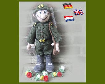May include: A crocheted toy soldier wearing a green uniform and a hat with a peace sign. The soldier is holding a brown bag and standing on a brick surface. There are small crocheted flowers on the ground in front of the soldier.