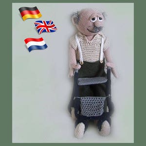 May include: A crocheted doll with grey hair and a moustache, wearing a white shirt with braces and brown trousers. The doll is holding a black walking frame with a grey seat.