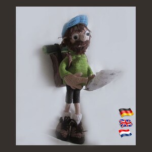 Può includere: Una bambola all'uncinetto con un cappello blu, una camicia verde, pantaloni marroni e stivali marroni. La bambola tiene una mappa e porta uno zaino marrone.