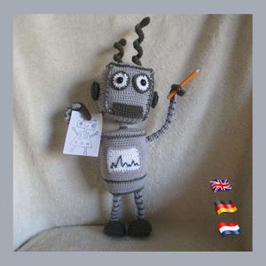 Op de afbeelding: Een grijs gehaakt robot speelgoed dat een vel papier vasthoudt met een tekening van een robot erop. De robot houdt een potlood in zijn rechterhand. De robot heeft een grijs lichaam met zwarte strepen op zijn benen en een grijs hoofd met zwarte ogen. De robot zit op een lichtgrijze ondergrond.