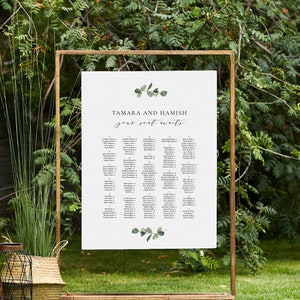 Könnte beinhalten: Eine weiße Hochzeitssitzordnung mit goldenem Rahmen, mit den Namen "Tamara und Hamish" und dem Text "Gässtesitzplan". Die Karte ist mit grünen Eukalyptusblättern verziert.