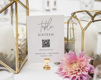 May include: A white table number card with the text "Table Sixteen" and a QR code. The card is on a gold stand and has a pink flower in front of it. The card also includes the names "Annalise + Brandon" and the date "September 1, 2022".